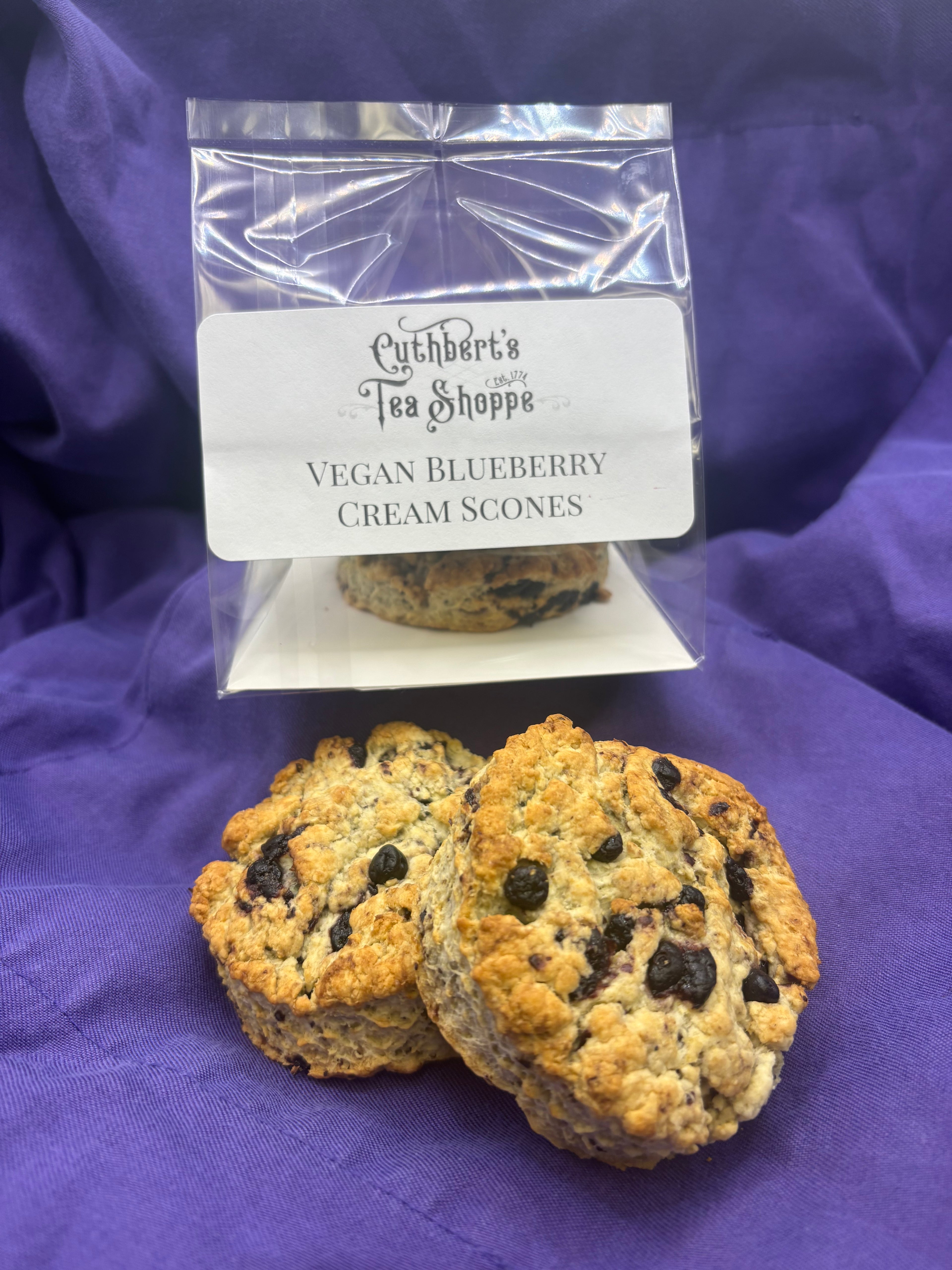 Packaged vegan blueberry scones with two individual scones set in front of it on purple fabric